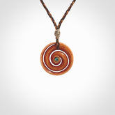 A hand carved and intricate koru pendant made for us by Yuri Terenyi. This is a beautiful little piece and is emblematic of the well known and loved Koru design. It is carved from stained bone in a hollowed, koru spiral shape with paua shell inlay in the middle of the koru. It is suspended from a gingernut brown/wapiti brown cord with a stardust floret and the necklace is adjustable.